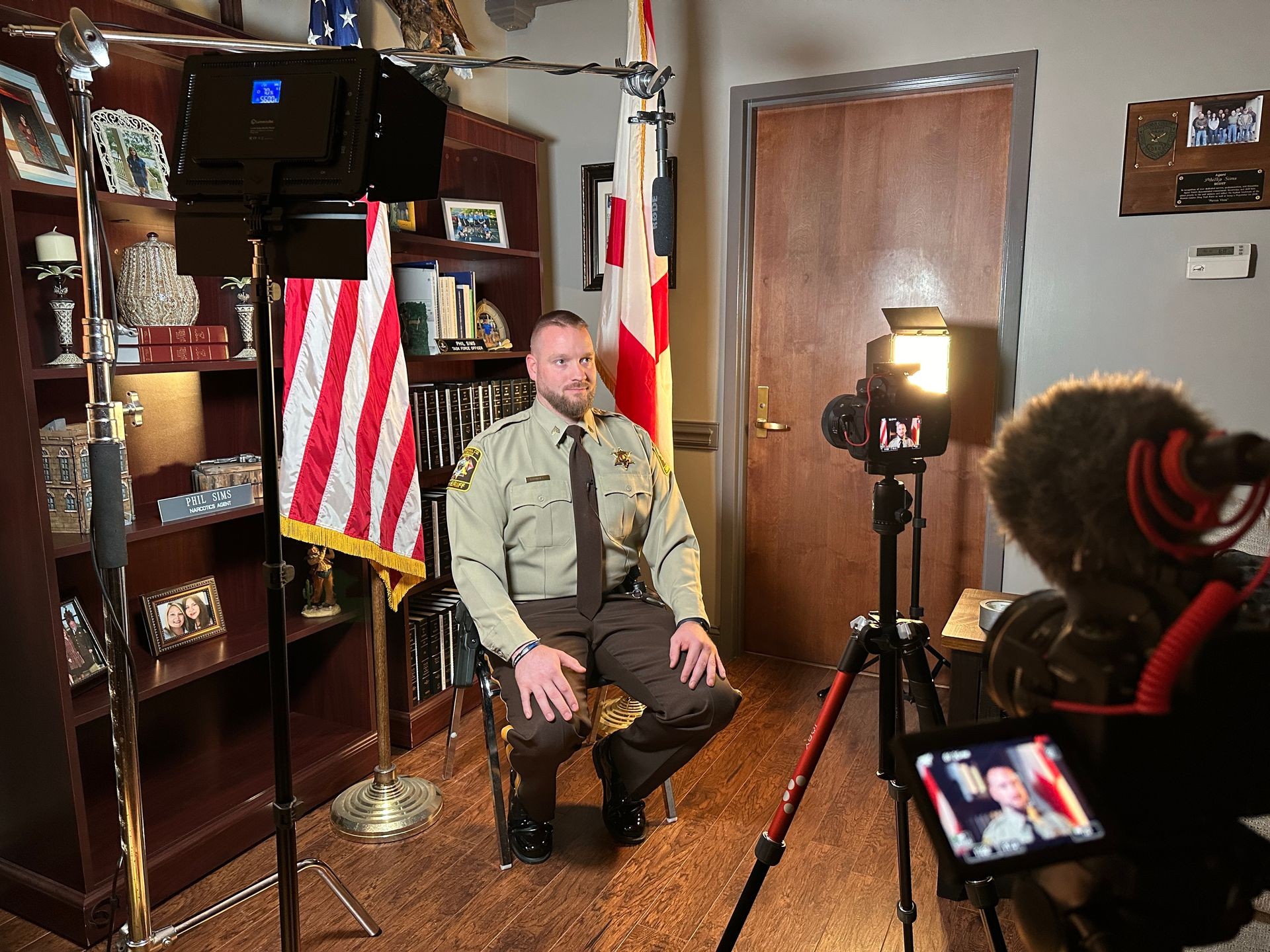 Man in sheriff uniform sits for an interview in an office with American flags and cameras recording.