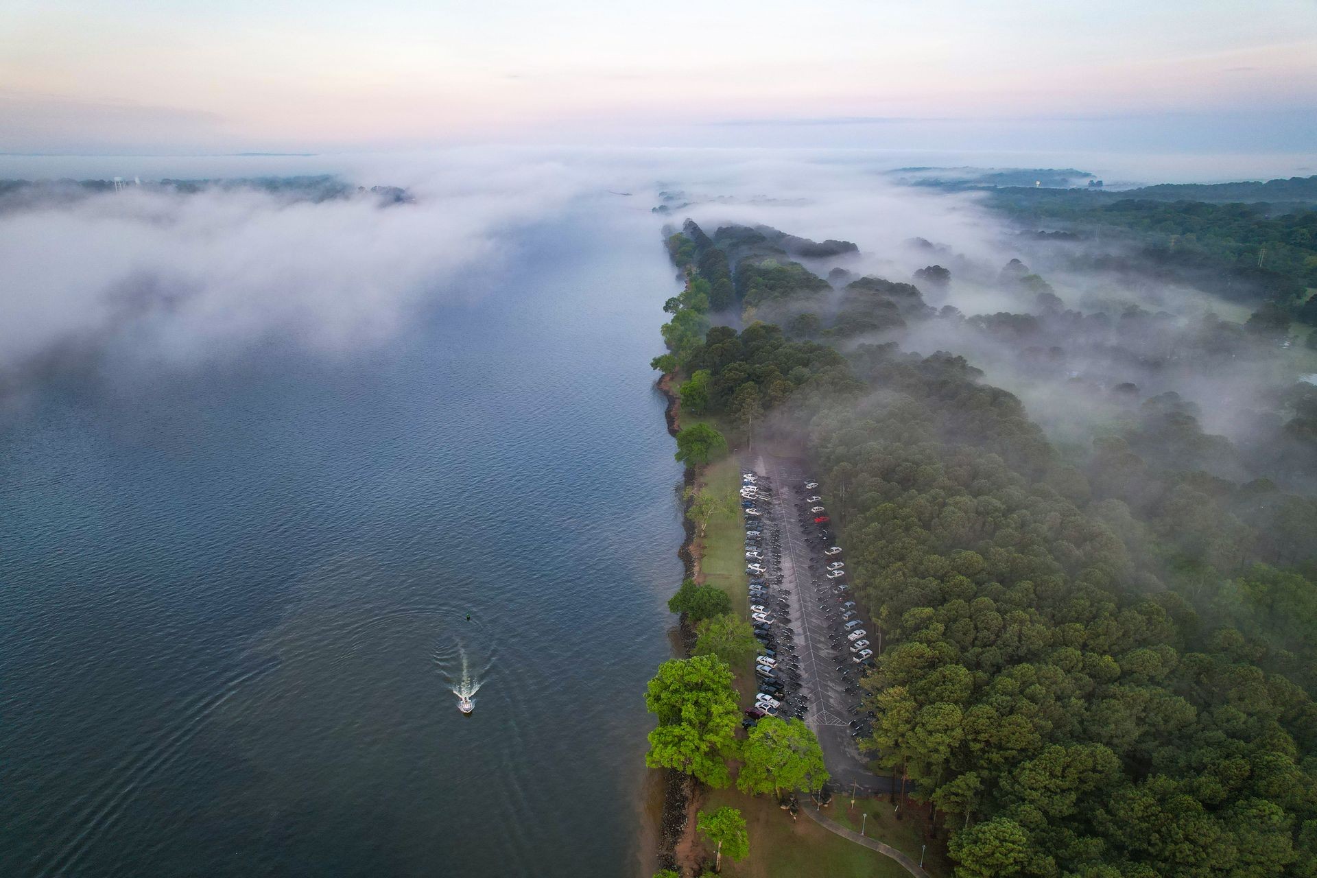 Aerial view of a river with surrounding foggy forest and a parking lot filled with cars.